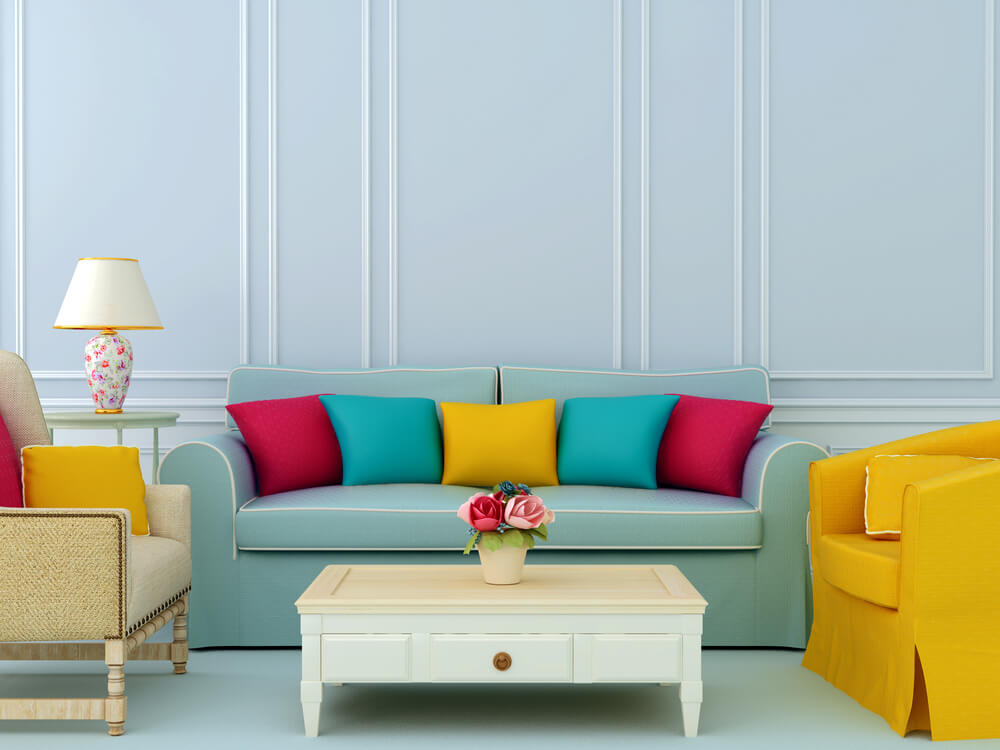 A decorated room. A couch, two chairs, an end table, and a coffee table. Bright and vibrant pillows on the light blue couch and one of the chairs being a brighter yellow color. 