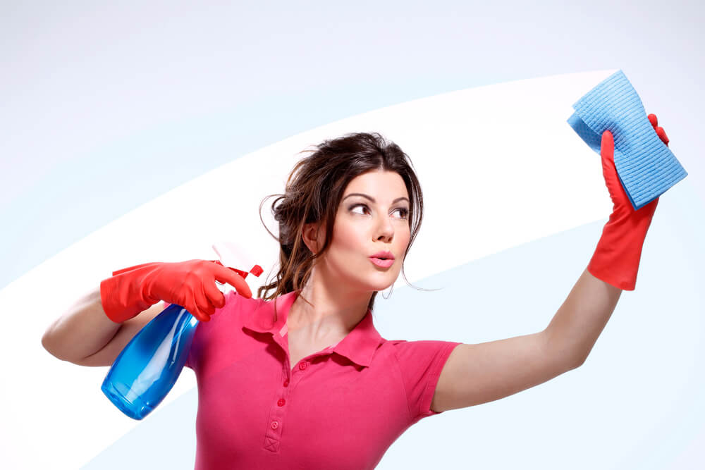 A woman holding a spray bottle of glass cleaner and wiping glass. Wearing a reddish pink polo and red gloves. 