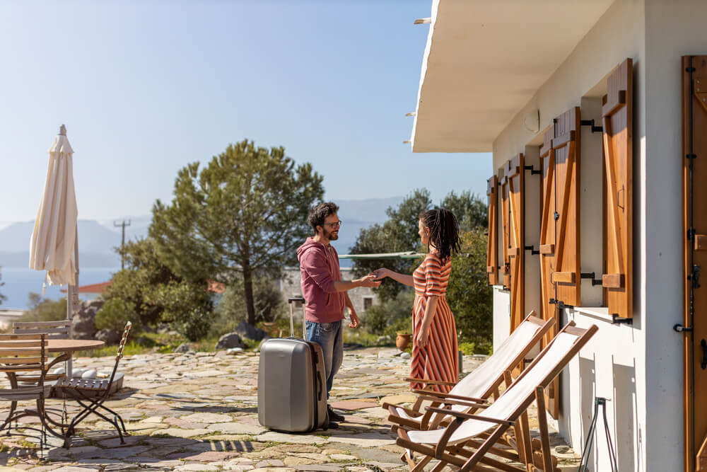 A picture of a guest checking in with luggage and a host greeting him. Mountains and water in the background. House is white with brown shutters. 